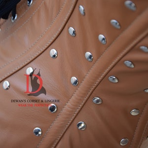 May include: Close-up of a brown leather corset with silver metal rivets. The corset has a seam down the center front and is made of a soft, supple leather. The text "DEWAN'S CORSET & LINGERIE - WEAR THE PERFECTION" is printed on the corset.
