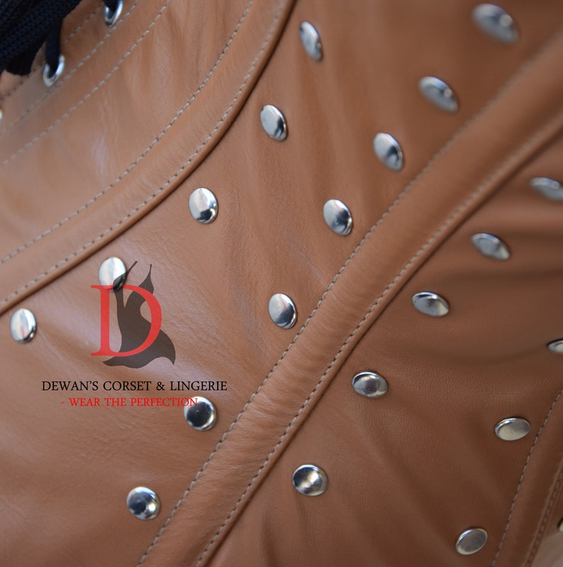 May include: Close-up of a brown leather corset with silver metal rivets. The corset has a seam down the center front and is made of a soft, supple leather. The text "DEWAN'S CORSET & LINGERIE - WEAR THE PERFECTION" is printed on the corset.
