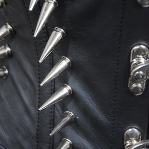 May include: Close-up of a black leather corset with silver metal spikes. The spikes are arranged in a row along the seam of the corset.