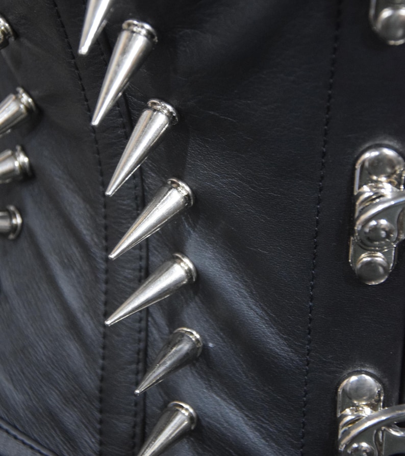 May include: Close-up of a black leather corset with silver metal spikes. The spikes are arranged in a row along the seam of the corset.
