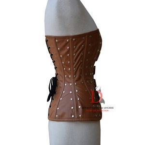 May include: A brown leather corset with silver studs and a black lace-up back. The corset has a strapless design and is made of a smooth, supple leather. "Dewan's Corset & Lingerie - Wear the Perfection" is printed on the bottom right corner of the corset.