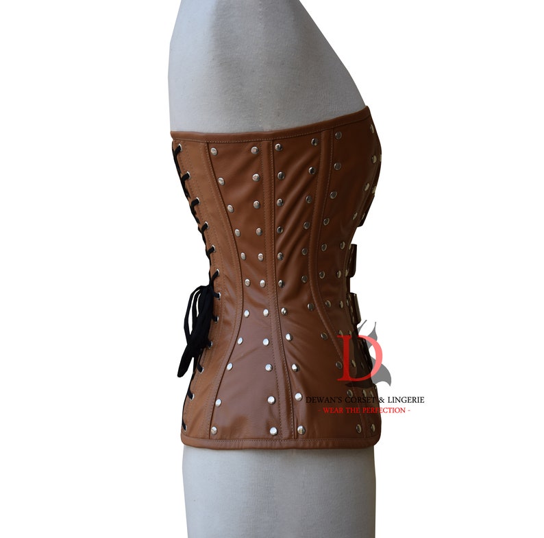 May include: A brown leather corset with silver studs and a black lace-up back. The corset has a strapless design and is made of a smooth, supple leather. "Dewan's Corset & Lingerie - Wear the Perfection" is printed on the bottom right corner of the corset.