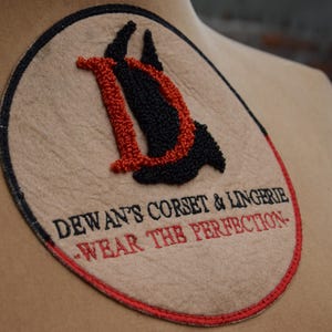 May include: A beige circular patch with red and black embroidery. The center features a stylized "D" with a red outline and black interior. The text "Dewant's Corset & Lingerie - Wear the Perfection" is embroidered in red around the "D".