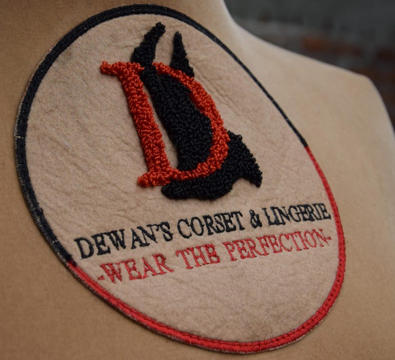 May include: A beige circular patch with red and black embroidery. The center features a stylized "D" with a red outline and black interior. The text "Dewant's Corset & Lingerie - Wear the Perfection" is embroidered in red around the "D".