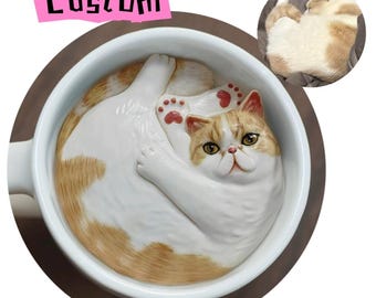 Puede incluir: Una taza de café de cerámica blanca con un gato marrón y blanco acurrucado en su interior. El gato tiene ojos naranjas y almohadillas rosadas. La taza tiene un asa en el lado izquierdo.