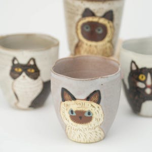 Könnte beinhalten: Eine Sammlung von Keramikbechern, die jeweils mit einer einzigartigen Katzenillustration verziert sind. Die Becher zeigen verschiedene Katzenrassen in Braun-, Schwarz- und Weißtönen. Ideal für Kaffee, Tee oder andere Getränke.