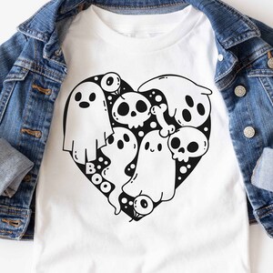 Puede incluir: Camiseta blanca con un gráfico en blanco y negro de fantasmas y calaveras en forma de corazón. El texto "BOO" está escrito debajo del corazón.