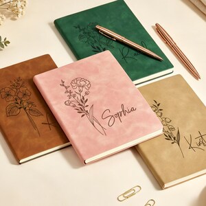 May include: Four personalized notebooks in brown, green, and pink with floral designs and names. Two rose gold pens and gold paper clips are also present. A cup of coffee sits on a white saucer.