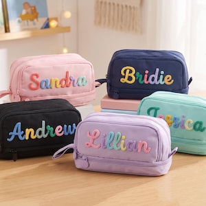 Op de afbeelding: Vijf rechthoekige etuis in roze, marineblauw, zwart, lavendel en lichtgroen. Elk etui heeft een ritssluiting en een handvat aan de zijkant. De voorzijde is gepersonaliseerd met een naam in kleurrijke, gestructureerde en verhoogde letters.
