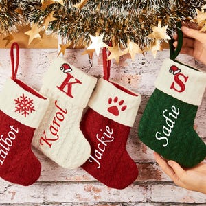 May include: Four Christmas stockings in red, white, and green hang from a garland. Each stocking is personalized with a name or initial in a script font. One stocking has a snowflake, another a paw print.