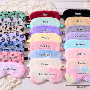 May include: A selection of plush sleep masks in a variety of colours and patterns, including leopard print and rainbow designs. The masks are arranged on a white surface, with labels indicating the colour of each mask. The masks measure 20cm wide and 10cm high.