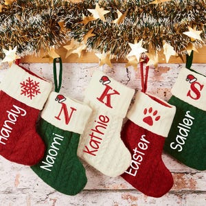 May include: Five Christmas stockings in red, green, and cream colors hang from a wooden beam. Each stocking is personalized with a name or initial in white, and some feature festive designs like a snowflake or paw print.