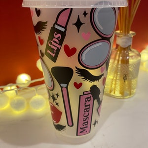 May include: A clear plastic tumbler with a pink, white and black design featuring makeup-themed illustrations, including lipstick, mascara, eyelashes and a makeup brush. The text "Lips" and "Mascara" are visible on the tumbler.