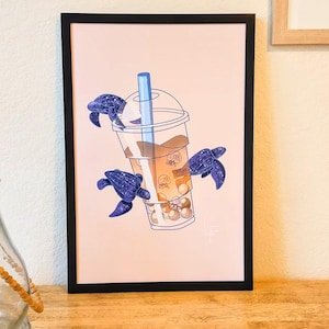 May include: A framed illustration of a boba tea with a straw and four leatherback sea turtles swimming around it. The background is a light peach color.