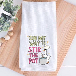 May include: White waffle weave kitchen towel with a cartoon goose wearing a chef's hat and stirring a pot. The text reads "On my way to stir the pot."