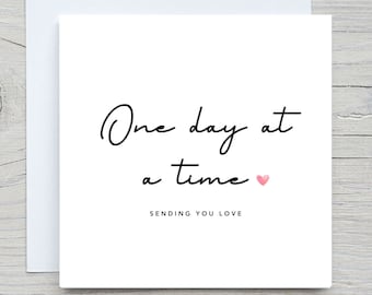 May include: A white greeting card with the words "One day at a time" in black script, a small pink heart, and the phrase "Sending you love" in a smaller font. The card is on top of a white envelope.