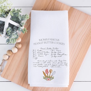 May include: White waffle-weave kitchen towel with the text "MOMS FAMOUS PEANUT BUTTER COOKIES" and a handwritten recipe. The towel features a floral and utensil design at the bottom, resting on a wooden cutting board.