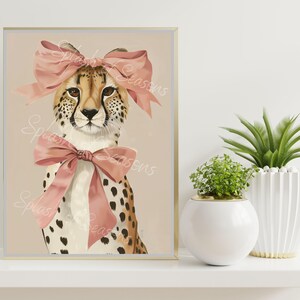 May include: A framed art print featuring a cheetah with a pink bow on its head and another around its neck. The cheetah has a tan coat with black spots. The background is a light beige color.