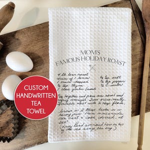 May include: White tea towel with a handwritten recipe for "Mom's Famous Holiday Roast" on it. The recipe includes ingredients like lean roast, lemon, onion, thyme, garlic, salt, pepper, and water. The text also includes instructions for preparing the roast, such as marinating overnight and browning in butter. The tea towel is on a wooden surface with a rolling pin, cookie cutters, and a wooden spoon.