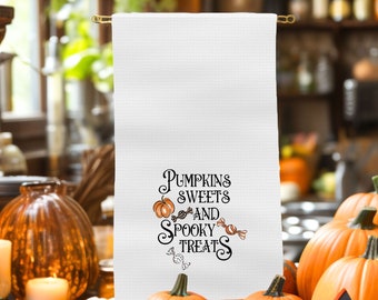 May include: White Halloween-themed kitchen towel with the words "Pumpkins, Sweets, and Spooky Treats" in black script. The design includes orange pumpkins and candy illustrations. The towel is hanging on a gold-colored rod, with pumpkins in the foreground.