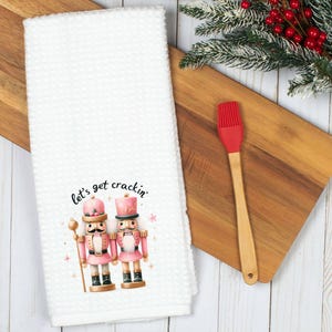 May include: White kitchen towel with a pink and gold nutcracker couple design. The text "let's get crackin'" is printed above the nutcrackers.