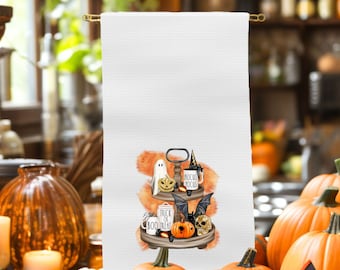 May include: A white kitchen towel with a Halloween-themed design. The design features a tiered tray with a ghost, a witch's hat, a mug that says "Hocus Pocus", a bat, a skull, and a pumpkin. The tray is decorated with orange and black accents. The text "Trick or Boo Treat" is written on the tray.