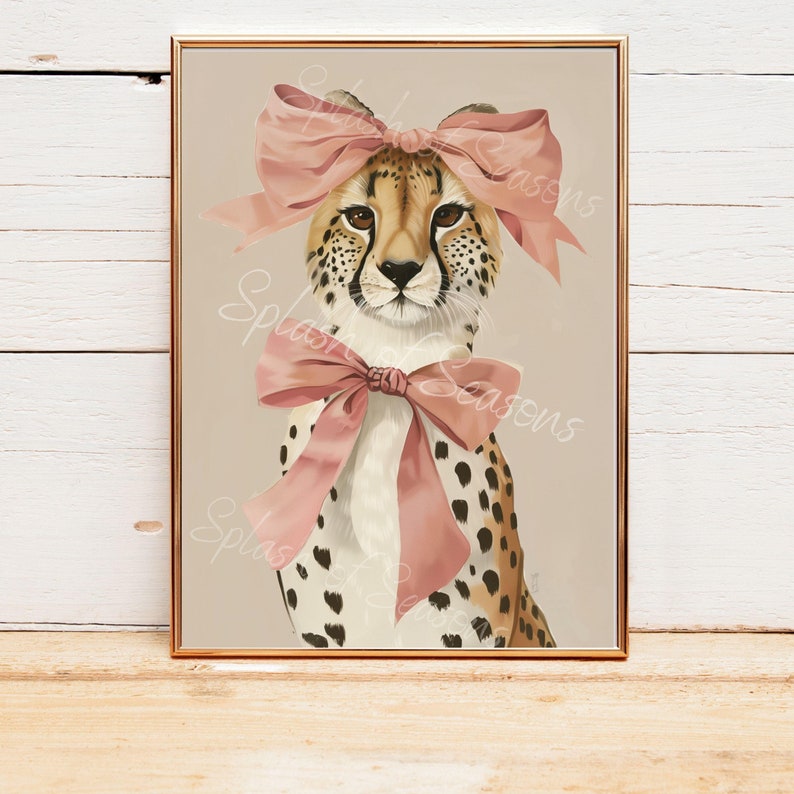 May include: A framed art print featuring a cheetah with a pink bow on its head and another around its neck. The cheetah has a tan and black spotted coat, with a light brown background. The print is in a gold frame and is displayed on a wooden surface.