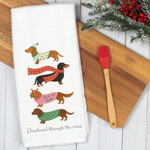 K&ouml;nnte beinhalten: Wei&szlig;es Waffel-K&uuml;chentuch mit vier Dackeln in festlicher Kleidung. Die Hunde tragen Pullover, Schals und Geweihe. Der Text "Deck the Halls" und "Dachshund through the snow" ist auf das Handtuch gedruckt. Ein roter Silikonpinsel und ein Holzschneidebrett befinden sich im Hintergrund.