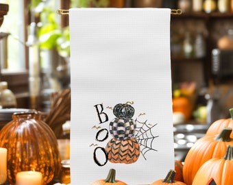 May include: A white kitchen towel with a black and gold "BOO" text and a stack of three pumpkins. The pumpkins are black and white checkered, black with a gold stem, and orange with a chevron pattern. A spiderweb is behind the pumpkins.