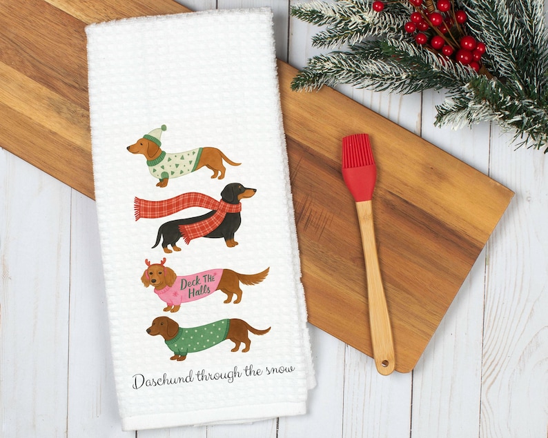 K&ouml;nnte beinhalten: Wei&szlig;es Waffel-K&uuml;chentuch mit vier Dackeln in festlicher Kleidung. Die Hunde tragen Pullover, Schals und Geweihe. Der Text "Deck the Halls" und "Dachshund through the snow" ist auf das Handtuch gedruckt. Ein roter Silikonpinsel und ein Holzschneidebrett befinden sich im Hintergrund.