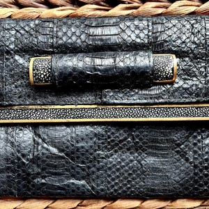 May include: Black clutch with a textured snakeskin pattern. The clutch features a handle with a gold trim and a contrasting speckled design. A gold-trimmed band with a speckled pattern accents the front. The clutch is set against a woven background.