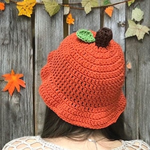Op de afbeelding: Een oranje gehaakte hoed in de vorm van een pompoen, met een groen blad en een bruine steel. Dit herfstaccessoire is handgemaakt. De hoed wordt van achteren getoond, met een houten hek en decoratieve bladeren op de achtergrond.
