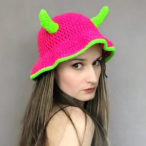 May include: A vibrant pink crochet hat with neon green edging and horns. The hat is a bucket style with a wide brim. The horns are neon green and attached to the top. The hat is handmade.