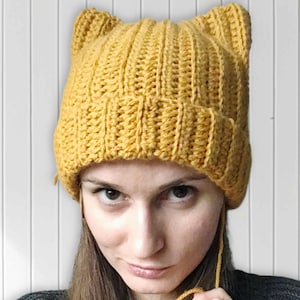 Peut inclure: Bonnet jaune moutarde en tricot avec des oreilles de chat. Le bonnet est fait d'un fil épais et texturé et présente un motif côtelé. Le chapeau est porté par une personne.