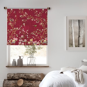 May include: A white bed with a white duvet cover and a beige throw blanket. The bed has a wooden frame and is sitting on a wooden floor. There is a red window blind with a floral pattern in the background.