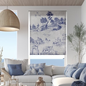 Puede incluir: Un sofá seccional blanco con almohadas y una manta azules. El sofá está en una sala de estar con una mesa de café de mimbre, una linterna y una ventana con vista al mar. La ventana tiene una persiana enrollable con estampado azul y blanco.