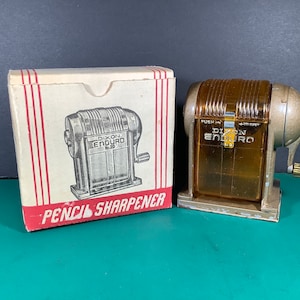 Puede incluir: Un sacapuntas vintage Dixon Enduro No. 30 en una caja de plástico marrón con base de metal. El sacapuntas está en su caja original, que es blanca con letras rojas que dicen "Pencil Sharpener".