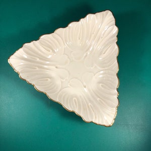 May include: A white triangular ceramic dish with a gold rim and a decorative floral pattern. The dish is on a teal background.