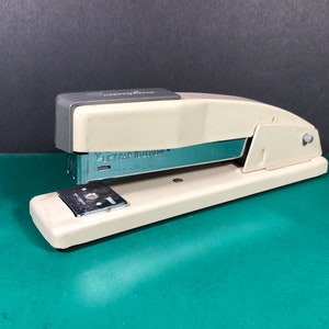 May include: A white and gray Swingline stapler with a silver staple remover. The stapler has a lift cap to hold the staples. The stapler is on a green surface.