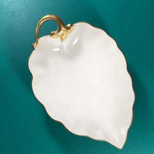 May include: A white porcelain leaf-shaped serving dish with a gold-tone handle and trim. The dish is on a teal background.