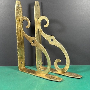 May include: A pair of L-shaped shelf brackets made of polished gold-toned metal. Each bracket features a hammered, textured surface and a decorative scrollwork support arm with a central circular cutout and pre-drilled holes for mounting.