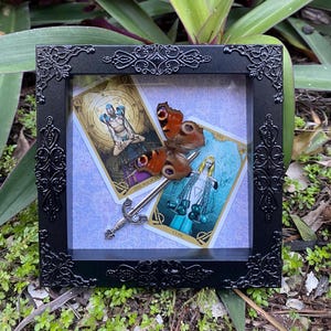 May include: A framed shadow box containing two tarot cards, "The Magician" and "Eight of Cups", a butterfly, and a silver sword. The background is a light blue with a white pattern.