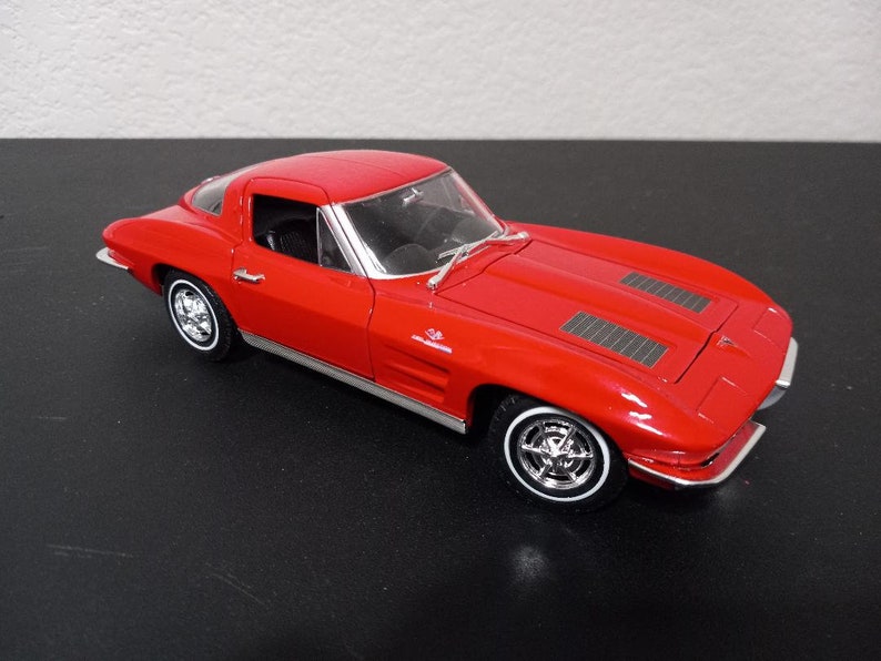 Puede incluir: Un coche modelo Chevrolet Corvette rojo de la d&eacute;cada de 1960 con interior negro y detalles cromados. El coche es un coup&eacute; con un cap&oacute; largo y una cubierta trasera corta. El coche est&aacute; sobre una superficie negra.