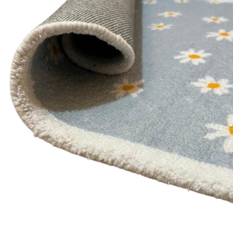 May include: A rolled-up rug featuring a light blue surface adorned with white daisies, each with a yellow centre. The rug has a white border and a grey backing.
