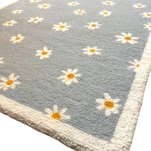 May include: A pale blue rug adorned with a daisy pattern. White daisies with yellow centres are spread across the rug's surface. The rug is edged with a white border.