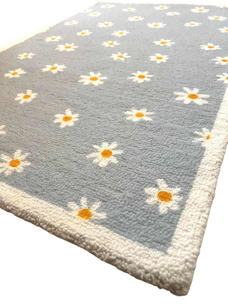 May include: A pale blue rug adorned with a daisy pattern. White daisies with yellow centres are spread across the rug's surface. The rug is edged with a white border.