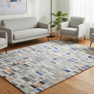 May include: A rectangular area rug featuring a geometric pattern of gray, white, and black blocks with interspersed accents of bright blue and soft orange. The rug is positioned on a light wood floor in front of a gray sofa and three gray armchairs.