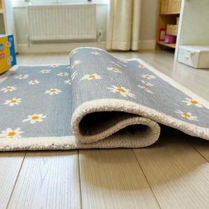 May include: A rolled-up, rectangular rug featuring a light grey backdrop adorned with a repeating pattern of white daisies with yellow centres. The rug has a white border and is on a light-coloured wooden floor.