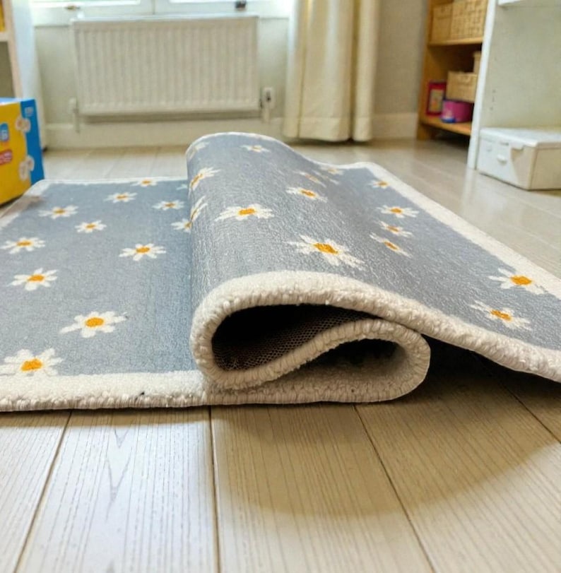May include: A rolled-up, rectangular rug featuring a light grey backdrop adorned with a repeating pattern of white daisies with yellow centres. The rug has a white border and is on a light-coloured wooden floor.
