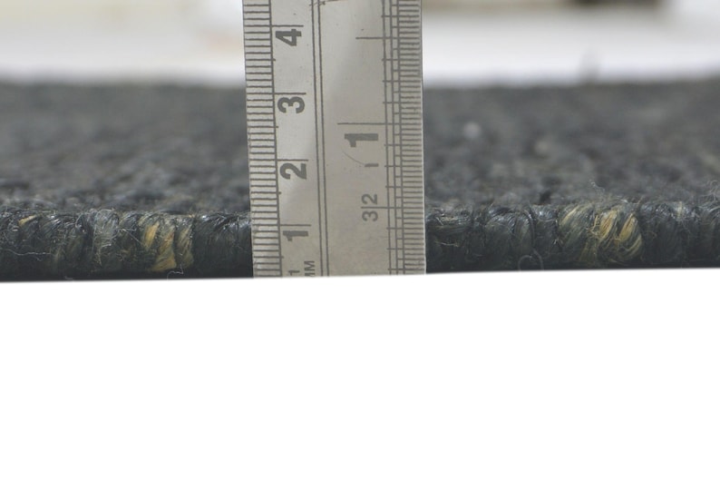 May include: Close-up of a dark gray rug with a textured surface. A metal ruler is positioned vertically, showing a thickness of approximately 0.5 inches. The rug's fibers have subtle variations in color, with hints of yellow.
